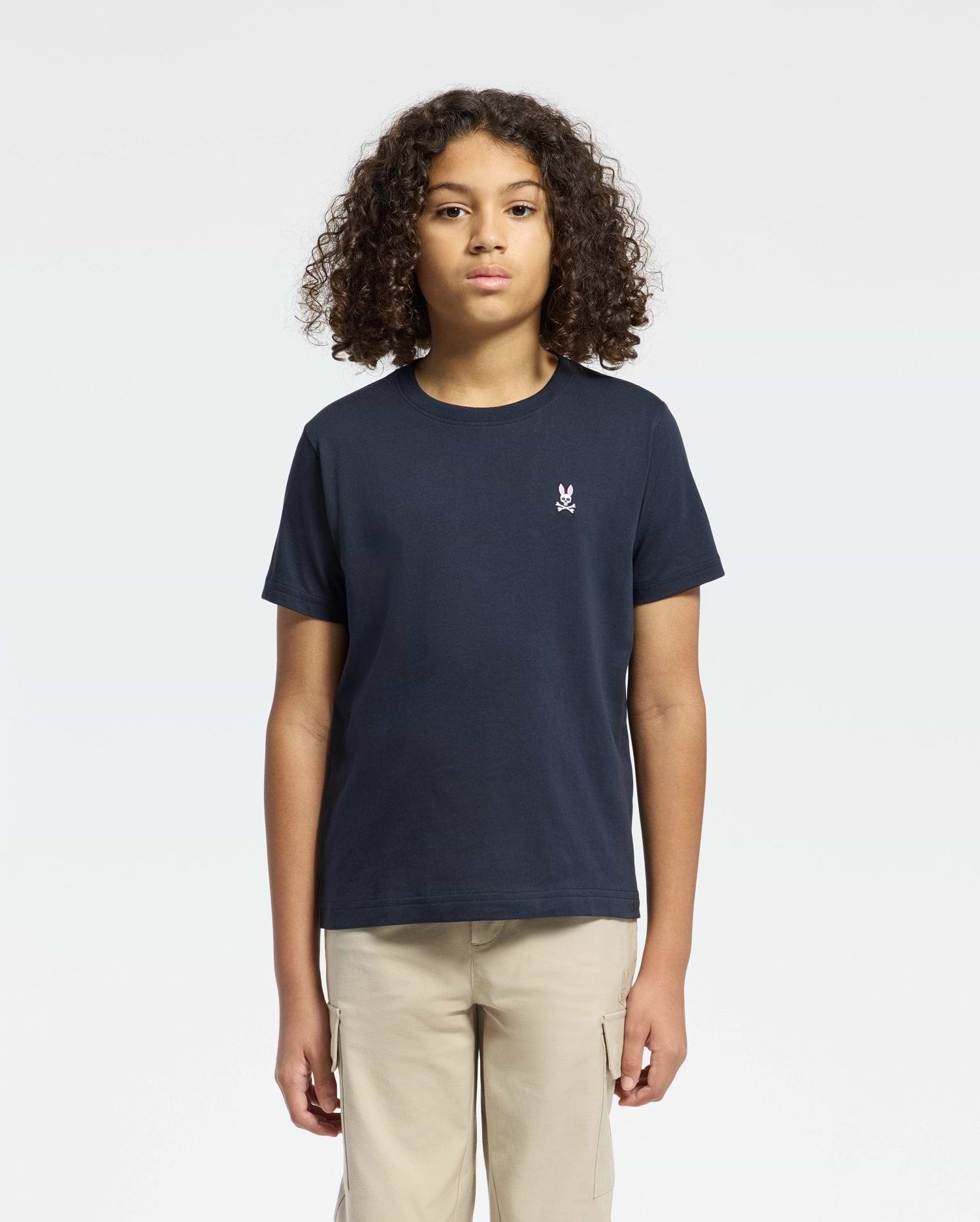 A curly-haired person wears a Psycho Bunny Kids Classic Crew Neck Tee in plain navy, made from 100% Pima cotton with a small chest embroidery. Paired with beige pants against a white background, they represent timeless basics.
