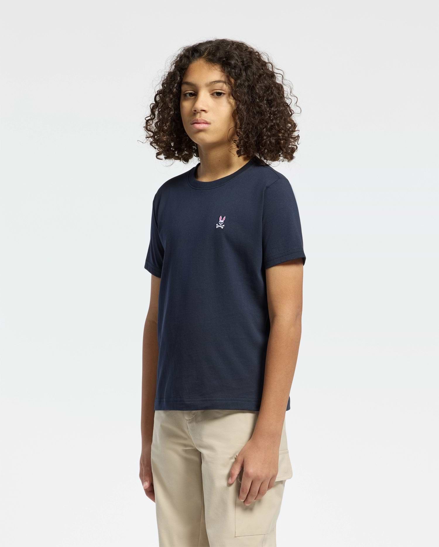 A young person with curly hair stands against a plain background, wearing a navy Kids Classic Crew Neck Tee by Psycho Bunny made from 100% Pima cotton and tan pants, exuding effortless style with their neutral expression and arms relaxed at their sides.