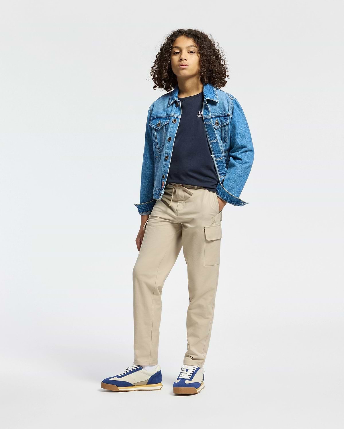 A person with curly hair wearing a classic denim jacket, a Psycho Bunny KIDS CLASSIC CREW NECK TEE in black, beige cargo pants, and blue and white sneakers stands confidently against a plain white background.