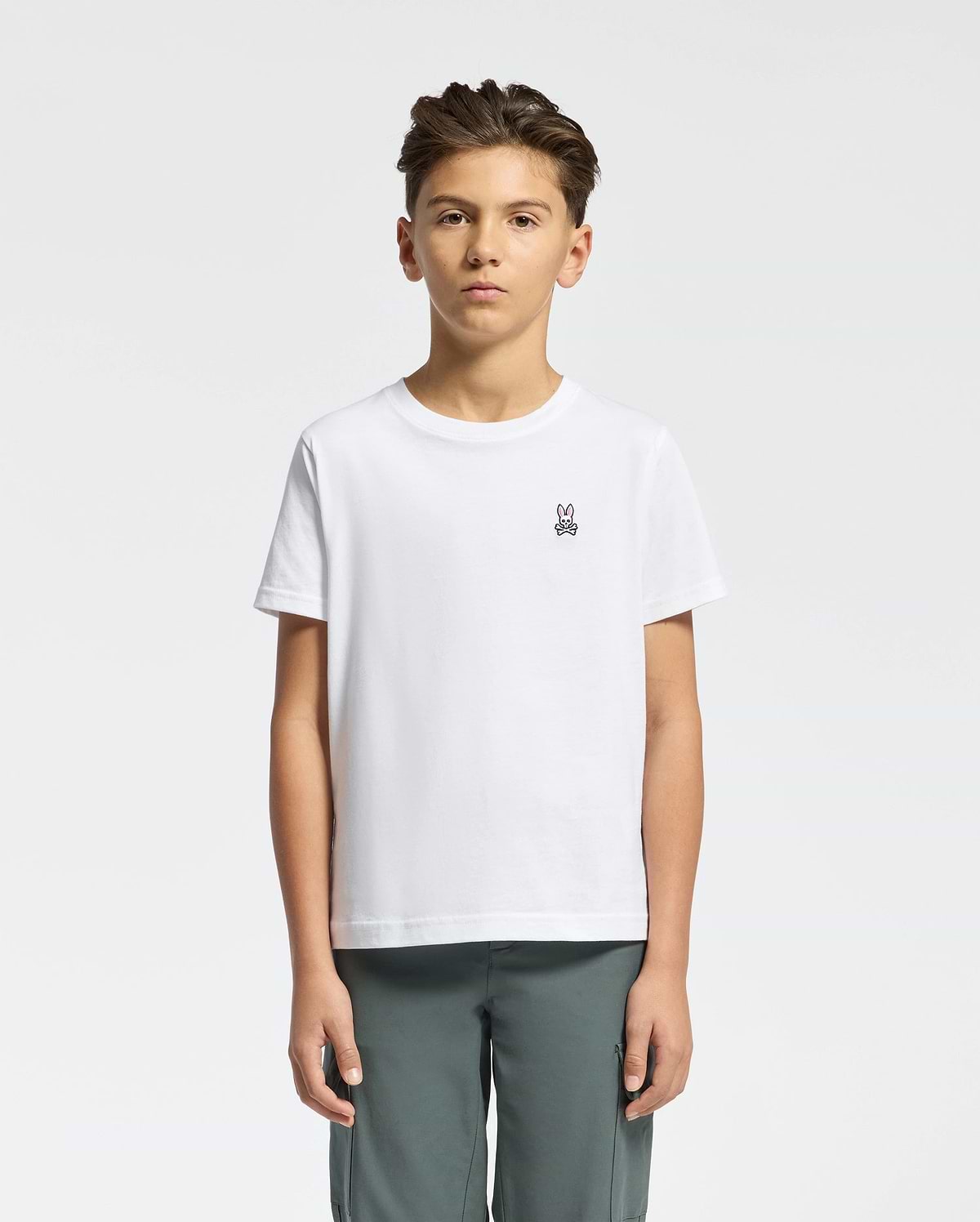 A young person with short dark hair, wearing a Psycho Bunny KIDS CLASSIC CREW NECK TEE - B0U014CRPC in crisp white 100% Pima cotton with the Classic Psycho Bunny embroidered logo, stands against a white background. They pair it with gray pants and maintain a neutral expression while facing forward.