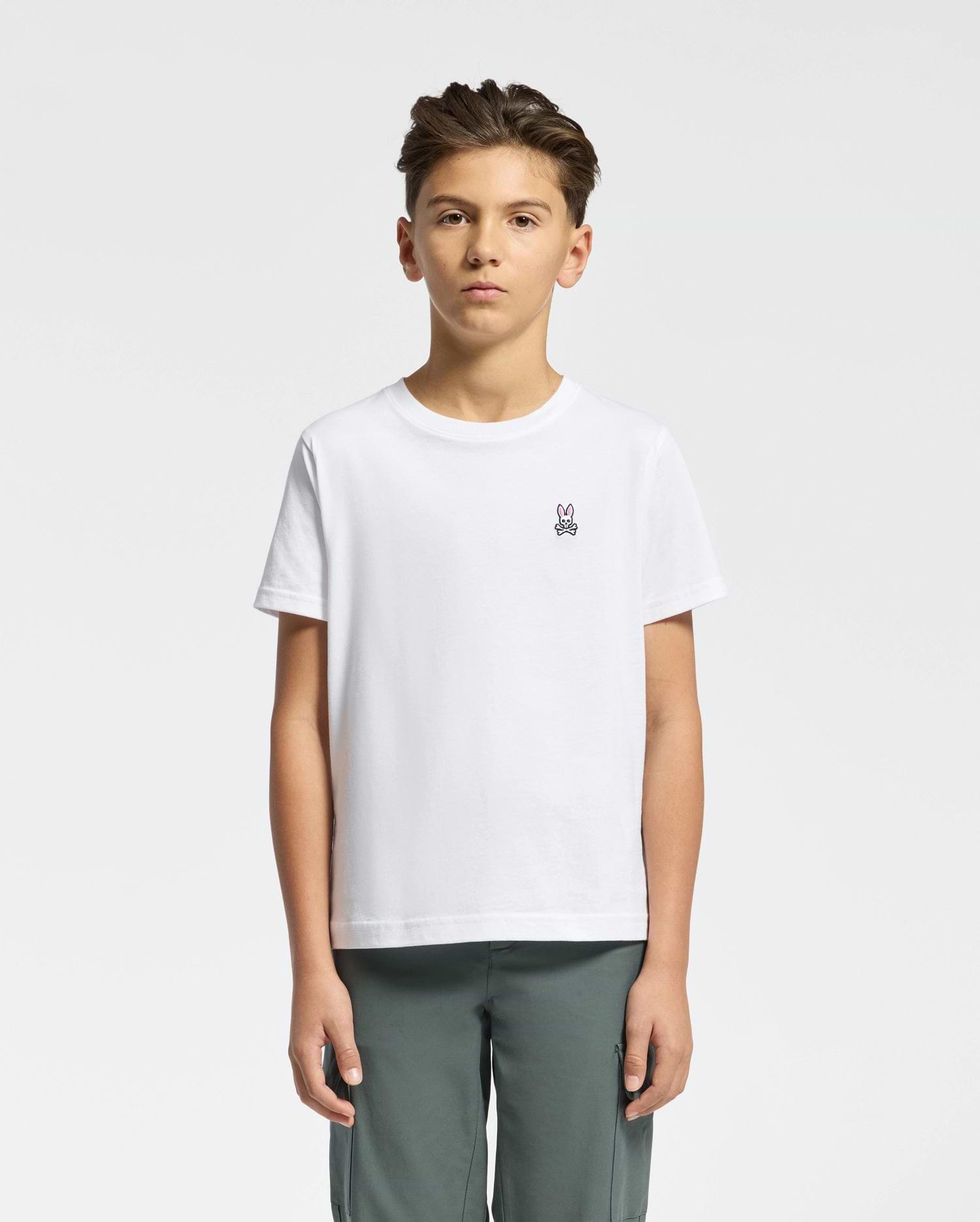 A young person with short dark hair, wearing a Psycho Bunny KIDS CLASSIC CREW NECK TEE - B0U014CRPC in crisp white 100% Pima cotton with the Classic Psycho Bunny embroidered logo, stands against a white background. They pair it with gray pants and maintain a neutral expression while facing forward.