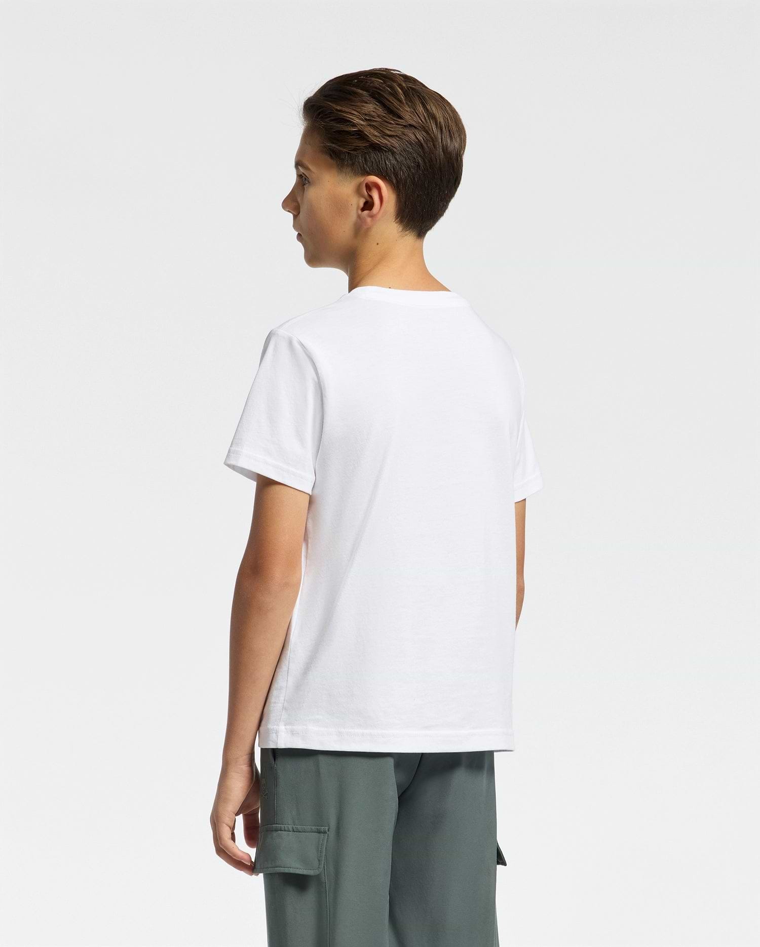 A young person with short brown hair stands sideways wearing Psycho Bunny's Kids Classic Crew Neck Tee in 100% Pima cotton and gray cargo pants. The plain white shirt showcases the classic embroidered Psycho Bunny logo on a neutral background.