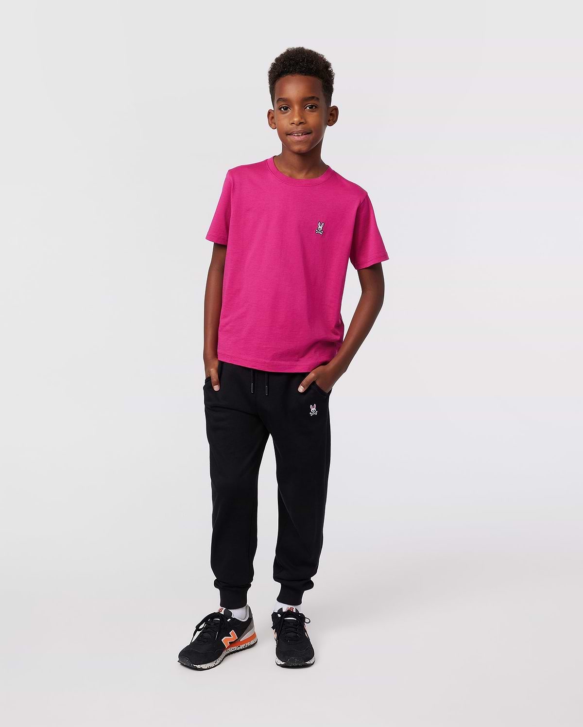 A young boy with short curly hair stands confidently against a plain background, wearing the Psycho Bunny KIDS CLASSIC CREW NECK TEE - B0U014CRPC in bright pink, paired with black joggers and black sneakers with orange accents.
