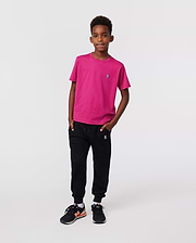 A young boy with short curly hair stands confidently against a plain background, wearing the Psycho Bunny KIDS CLASSIC CREW NECK TEE - B0U014CRPC in bright pink, paired with black joggers and black sneakers with orange accents.