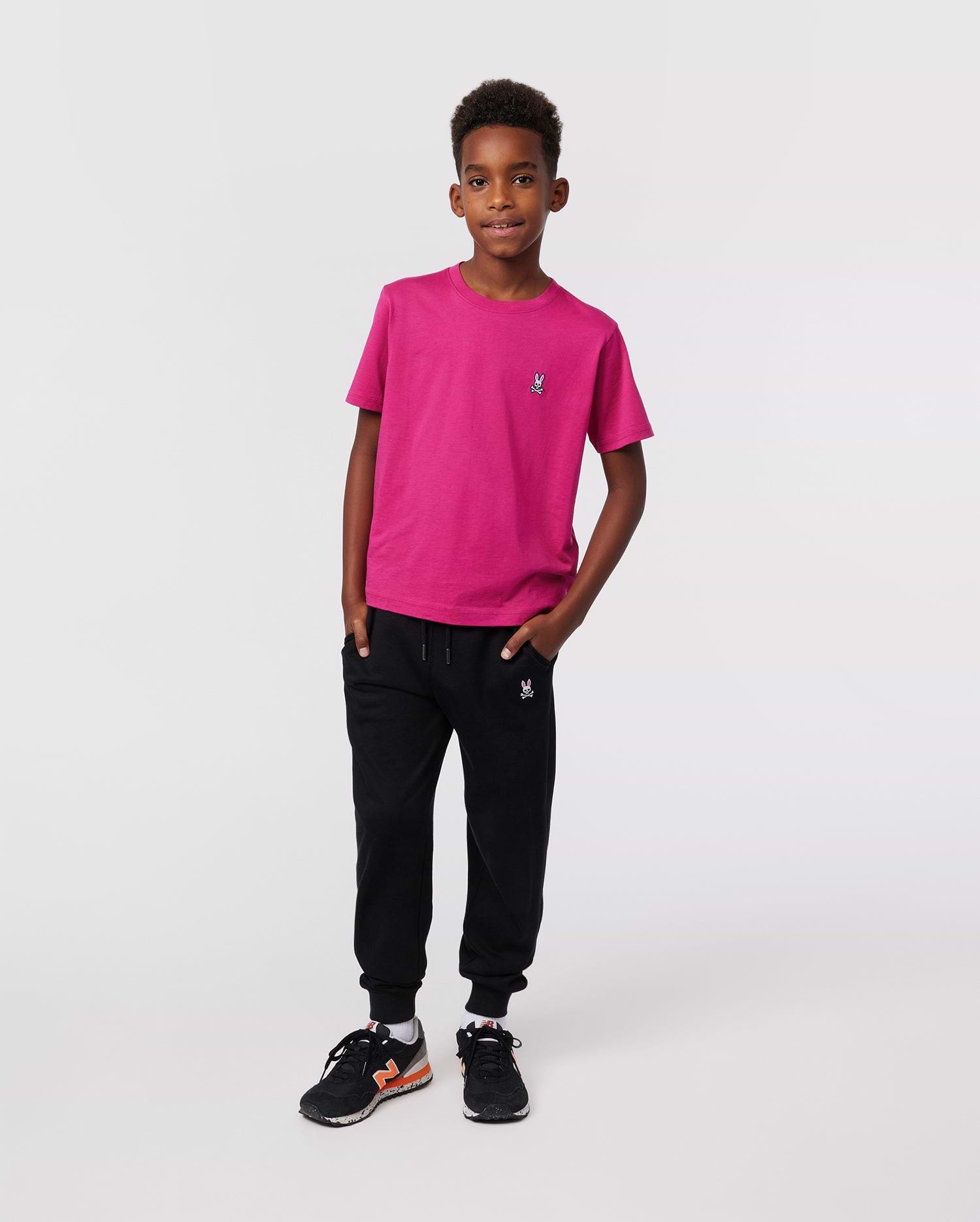 A young boy with short curly hair stands confidently against a plain background, wearing the Psycho Bunny KIDS CLASSIC CREW NECK TEE - B0U014CRPC in bright pink, paired with black joggers and black sneakers with orange accents.