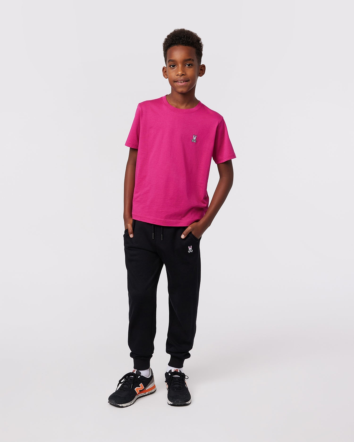 A young boy with short curly hair stands confidently against a plain background, wearing the Psycho Bunny KIDS CLASSIC CREW NECK TEE - B0U014CRPC in bright pink, paired with black joggers and black sneakers with orange accents.