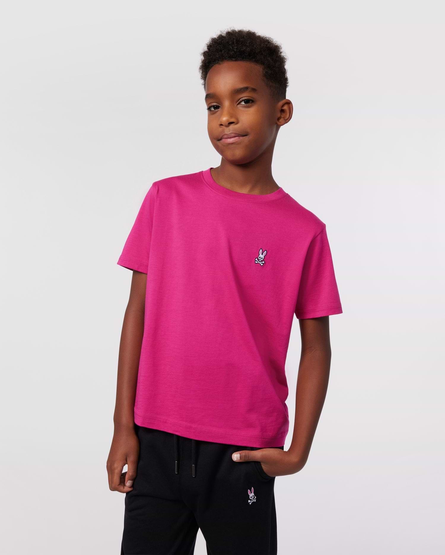 A boy stands confidently with one hand in his pocket, wearing Psycho Bunny’s KIDS CLASSIC CREW NECK TEE (B0U014CRPC) in premium Pima cotton with an embroidered logo and black pants, set against a plain light background.