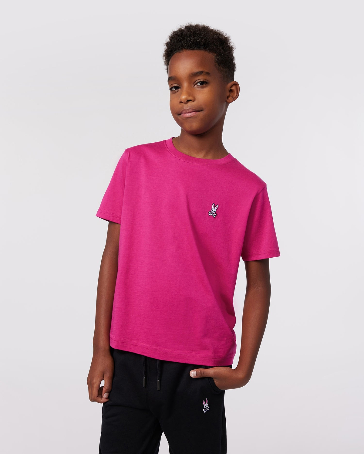 A boy stands confidently with one hand in his pocket, wearing Psycho Bunny’s KIDS CLASSIC CREW NECK TEE (B0U014CRPC) in premium Pima cotton with an embroidered logo and black pants, set against a plain light background.