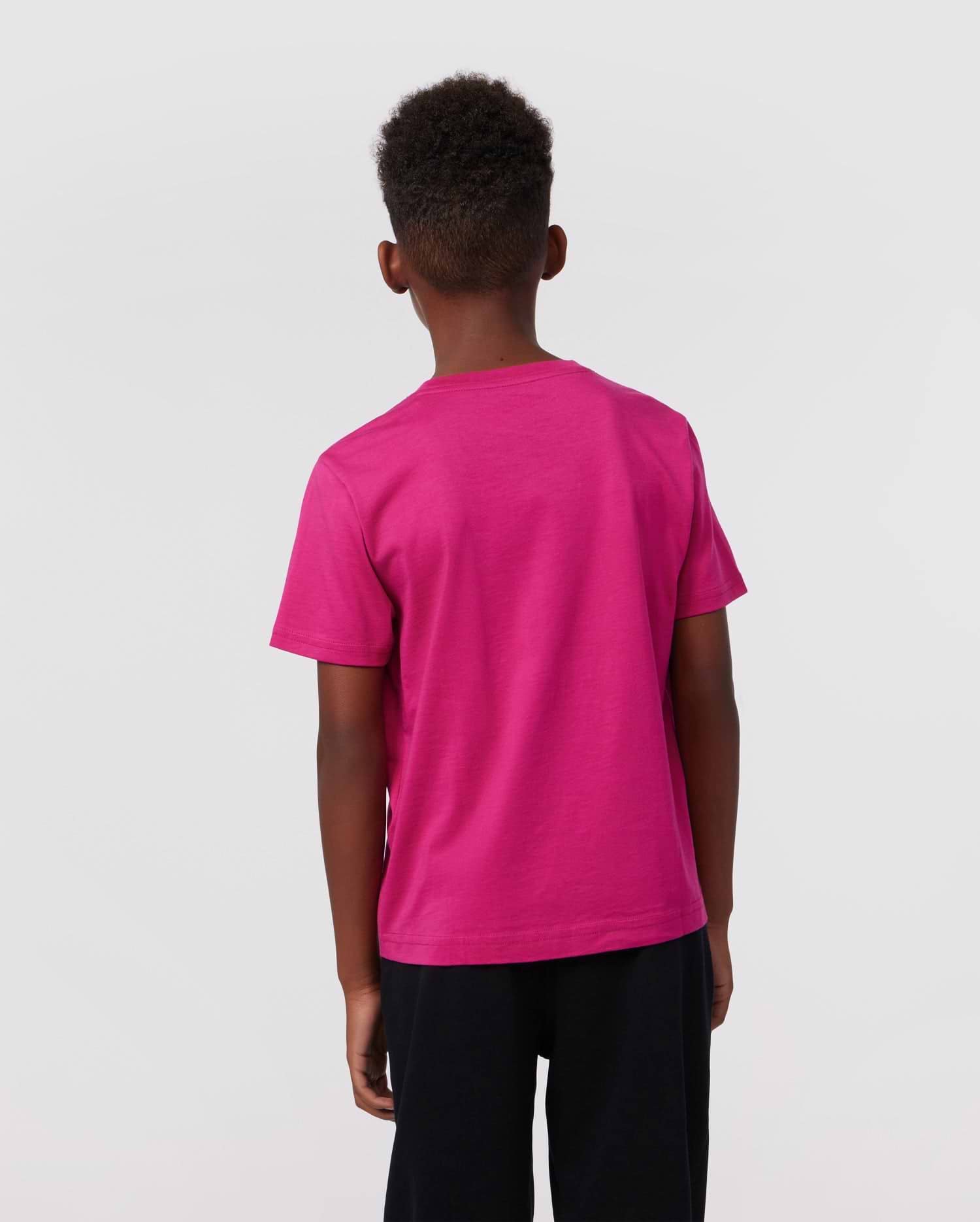 A young boy with short curly hair faces away from the camera, wearing the Psycho Bunny KIDS CLASSIC CREW NECK TEE - B0U014CRPC in vibrant pink and paired with black shorts, set against a plain light gray background.