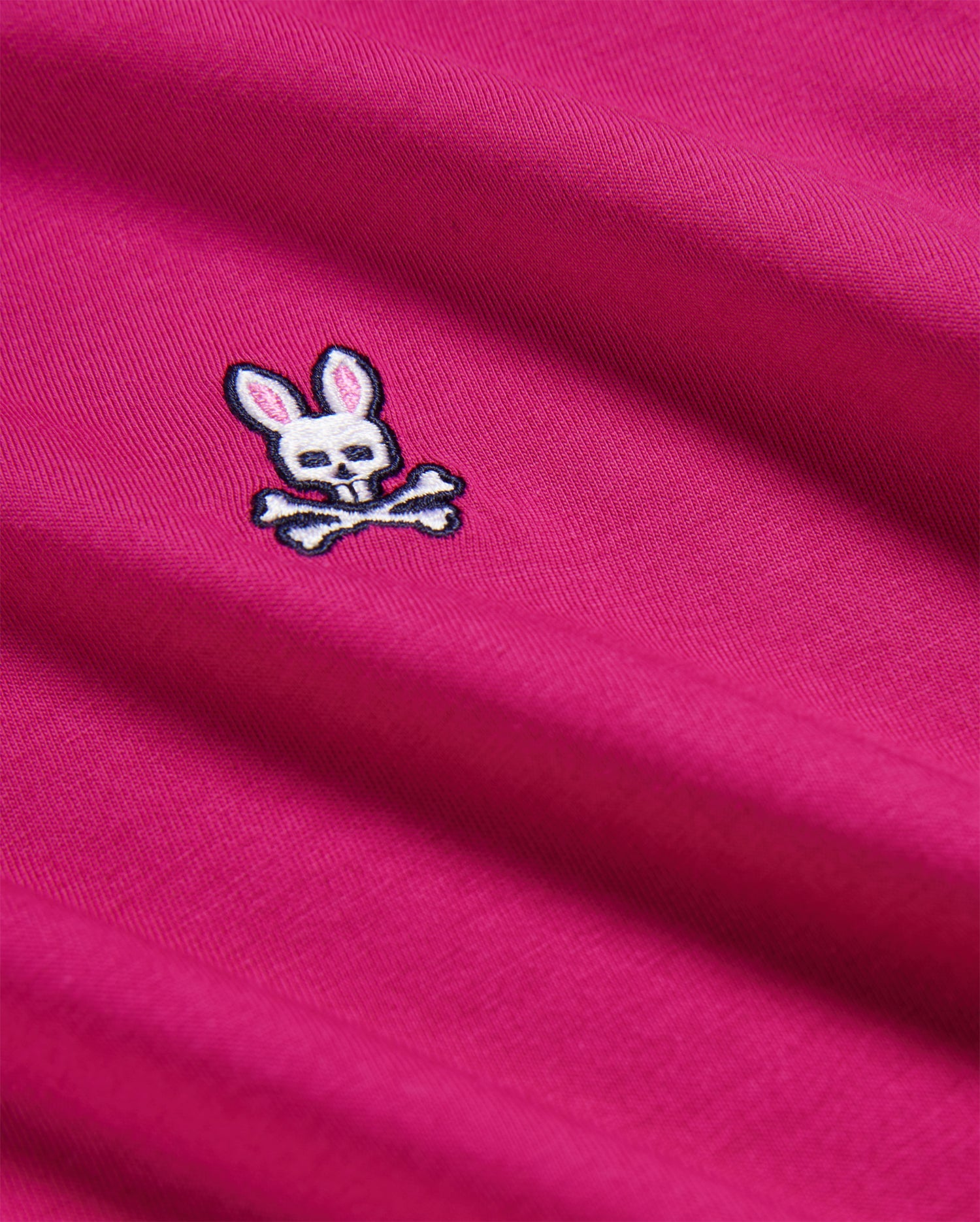 A close-up of the KIDS CLASSIC CREW NECK TEE - B0U014CRPC by Psycho Bunny in magenta premium Pima cotton, featuring an embroidered white bunny logo with pink ears and a skull face above crossed bones.