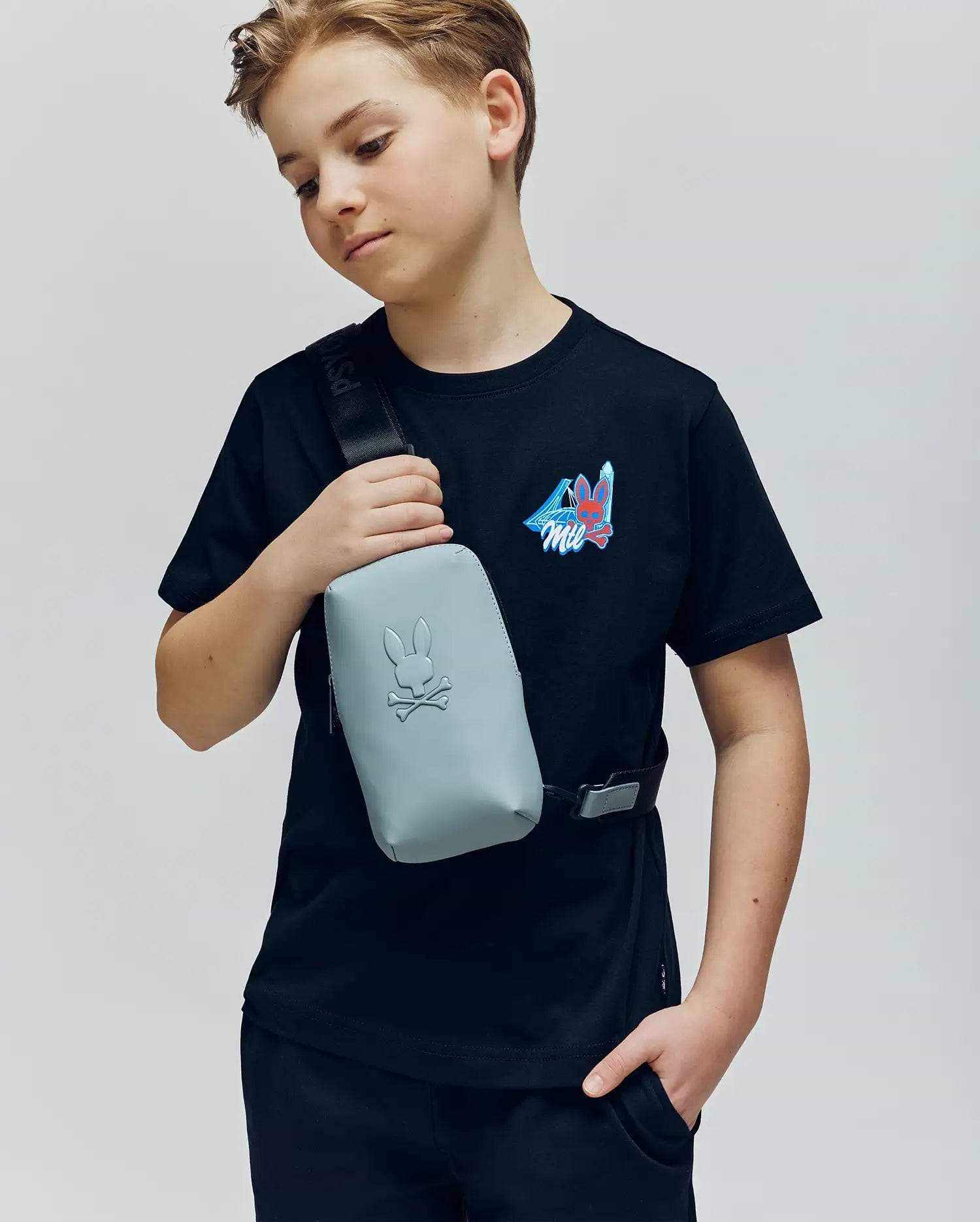 A boy in a Psycho Bunny KIDS MONTREAL TEE (B0U381F200)—black with a small colorful graphic—pairs it with a light gray crossbody bag, standing with one hand in his pocket. Made from 100% Pima cotton, it channels cool Montreal style.