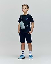 A young boy stands against a plain background wearing the Psycho Bunny KIDS MONTREAL TEE (B0U381F200), black shorts, blue sneakers, striped socks, and a gray crossbody bag. He looks straight ahead with hands in his pockets.