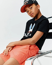 Black jersey t-shirt with large white Psycho Bunny skull logo, light blue ears, orange sunglasses. Paired with Kids Samos Hydrochromic Printed Swim Trunk in coral with small embroidered logo, and a black cap with orange brim.
