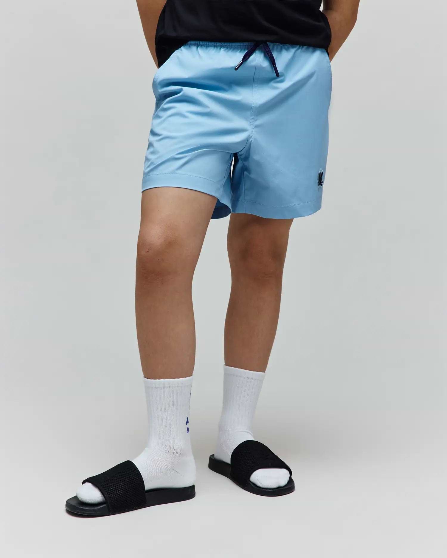 Kids Samos Hydrochromic Printed Swim Trunk in light blue, featuring a relaxed fit, smooth quick-dry fabric, elastic waistband with navy drawstring, and a small embroidered Psycho Bunny skull logo on the left leg.