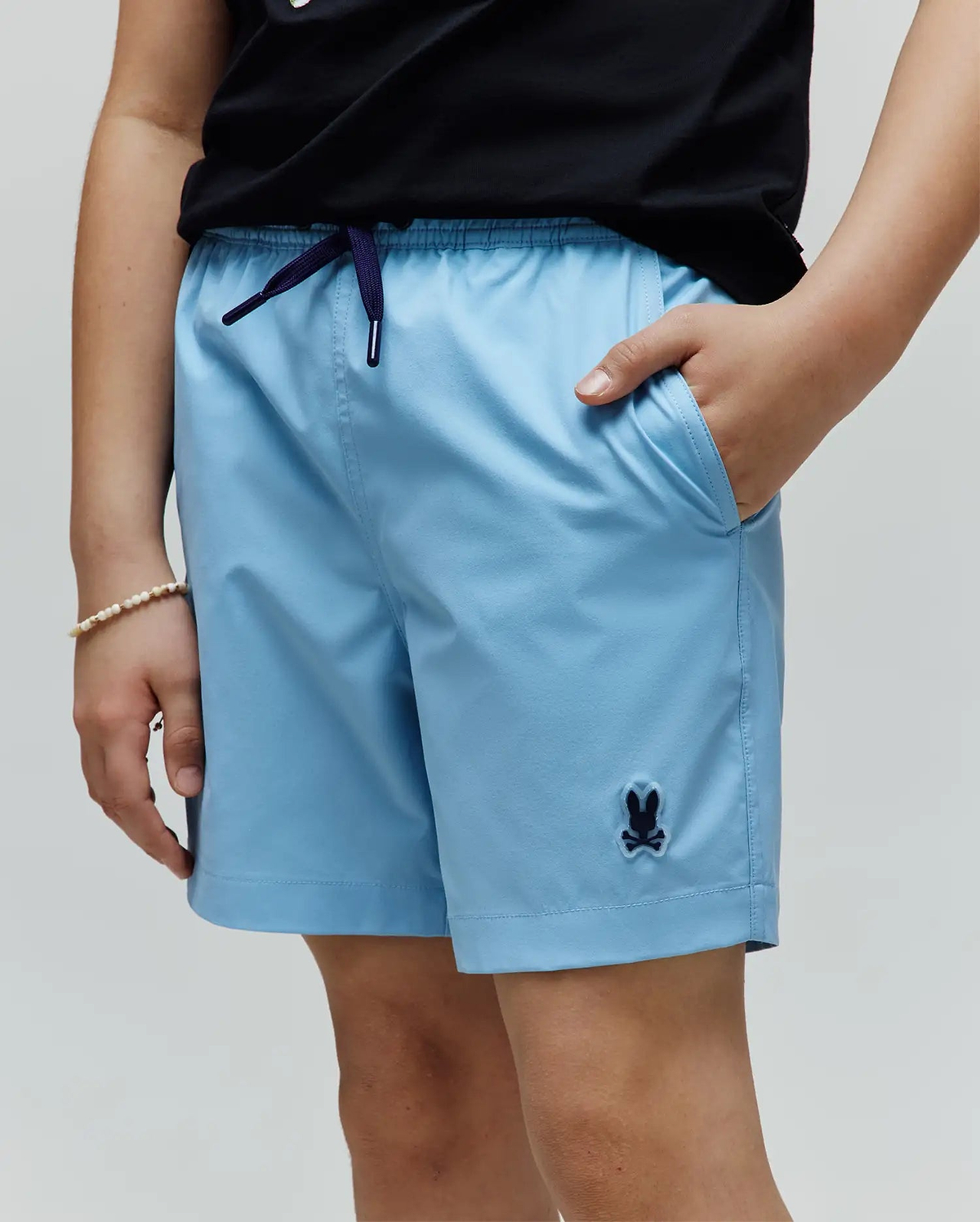 Kids Samos Hydrochromic Printed Swim Trunk in light blue, featuring a dark blue drawstring, side pockets, and a small embroidered Psycho Bunny skull logo on the left leg.