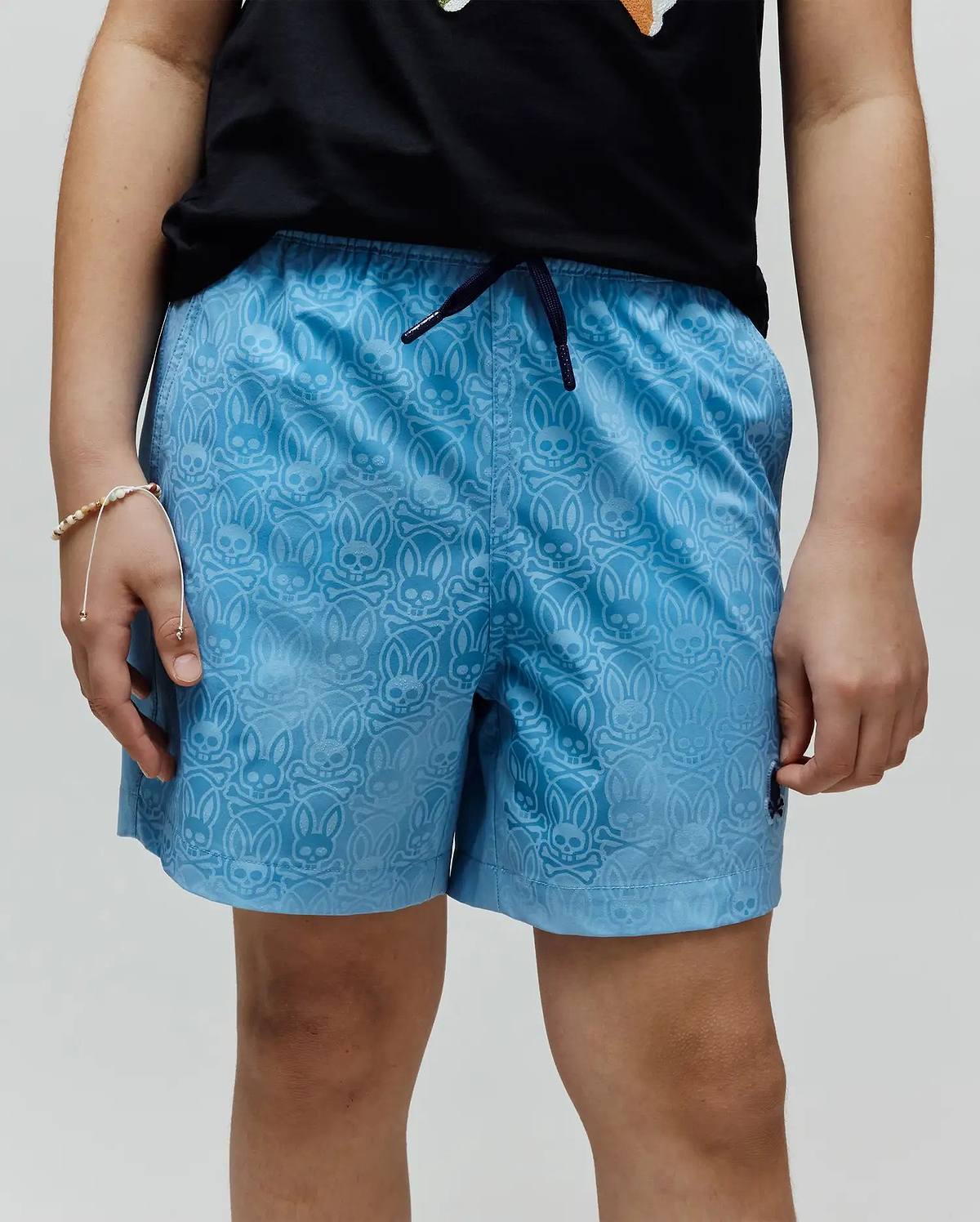 Kids Samos Hydrochromic Printed Swim Trunk in light blue, featuring an all-over tonal Psycho Bunny skull and crossbones pattern, dark blue drawstring, and small embroidered logo on the lower left leg.