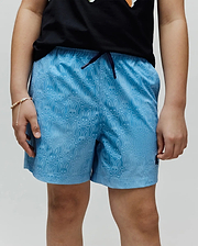 Kids Samos Hydrochromic Printed Swim Trunk in light blue, featuring an all-over tonal Psycho Bunny skull and crossbones pattern, dark blue drawstring, and small embroidered logo on the lower left leg.