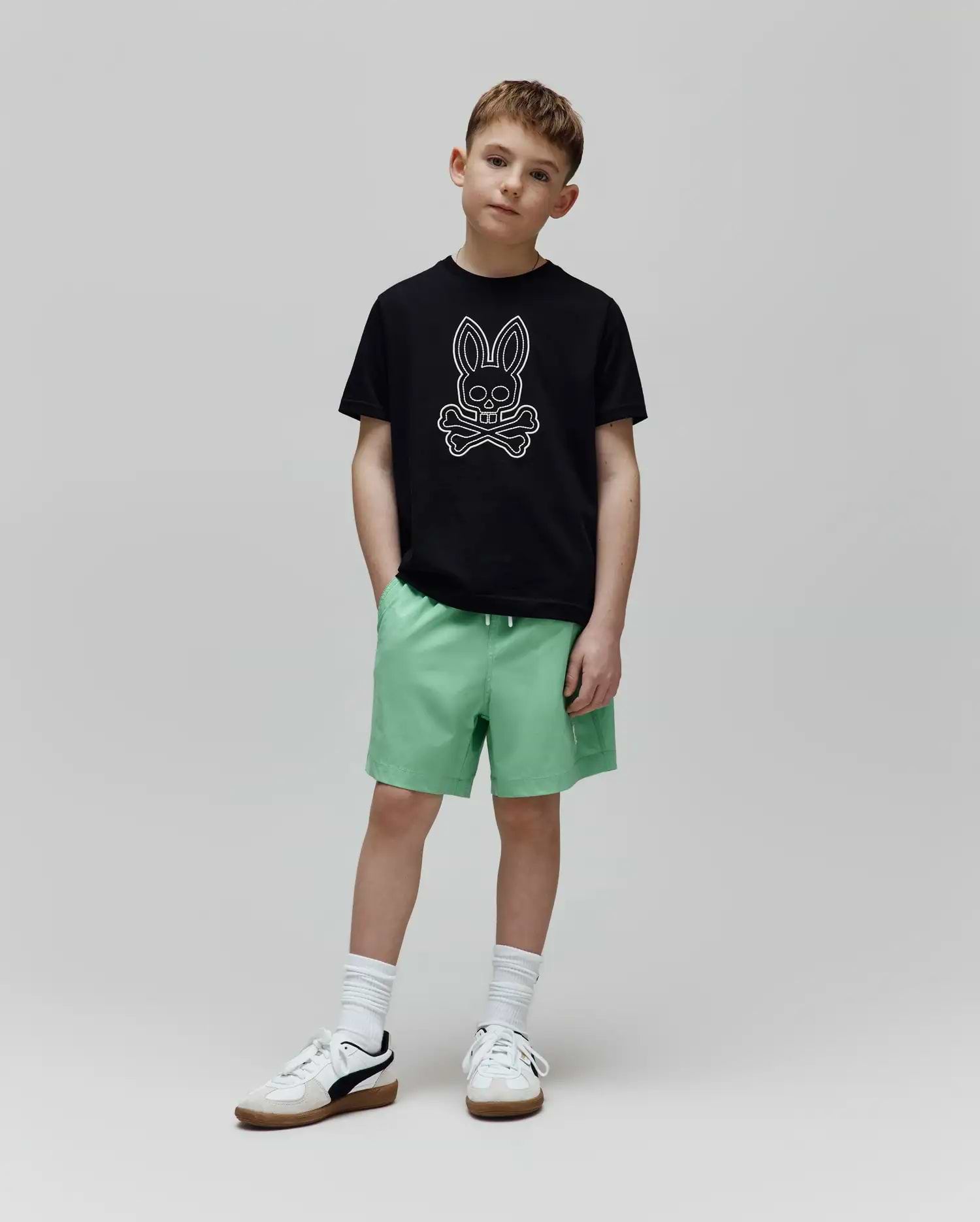 Black classic-fit jersey t-shirt with a large white outline Psycho Bunny skull logo on the chest, and light green Kids Samos Hydrochromic Printed Swim Trunk with a white drawstring.