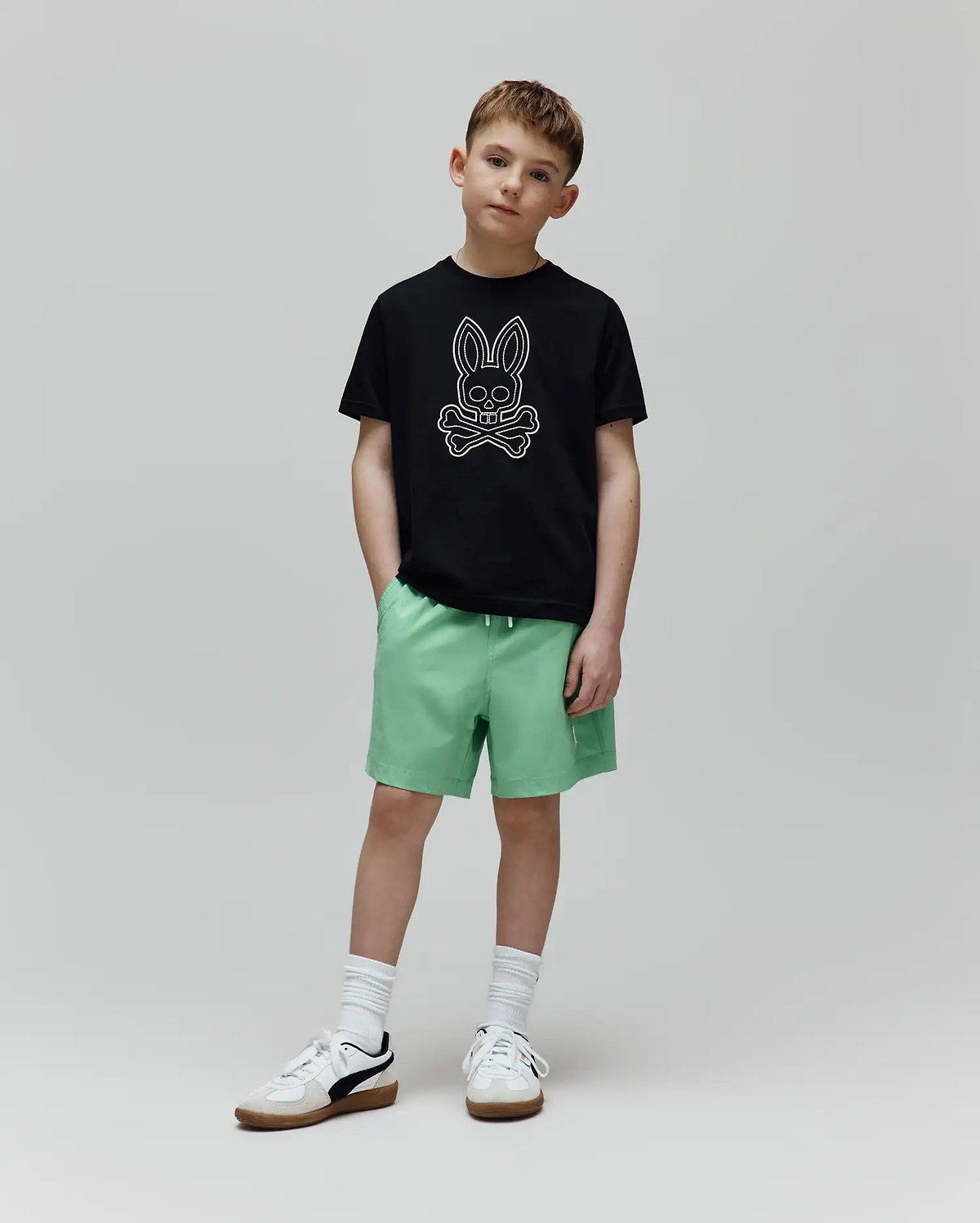 Black classic-fit jersey t-shirt with a large white outline Psycho Bunny skull logo on the chest, and light green Kids Samos Hydrochromic Printed Swim Trunk with a white drawstring.