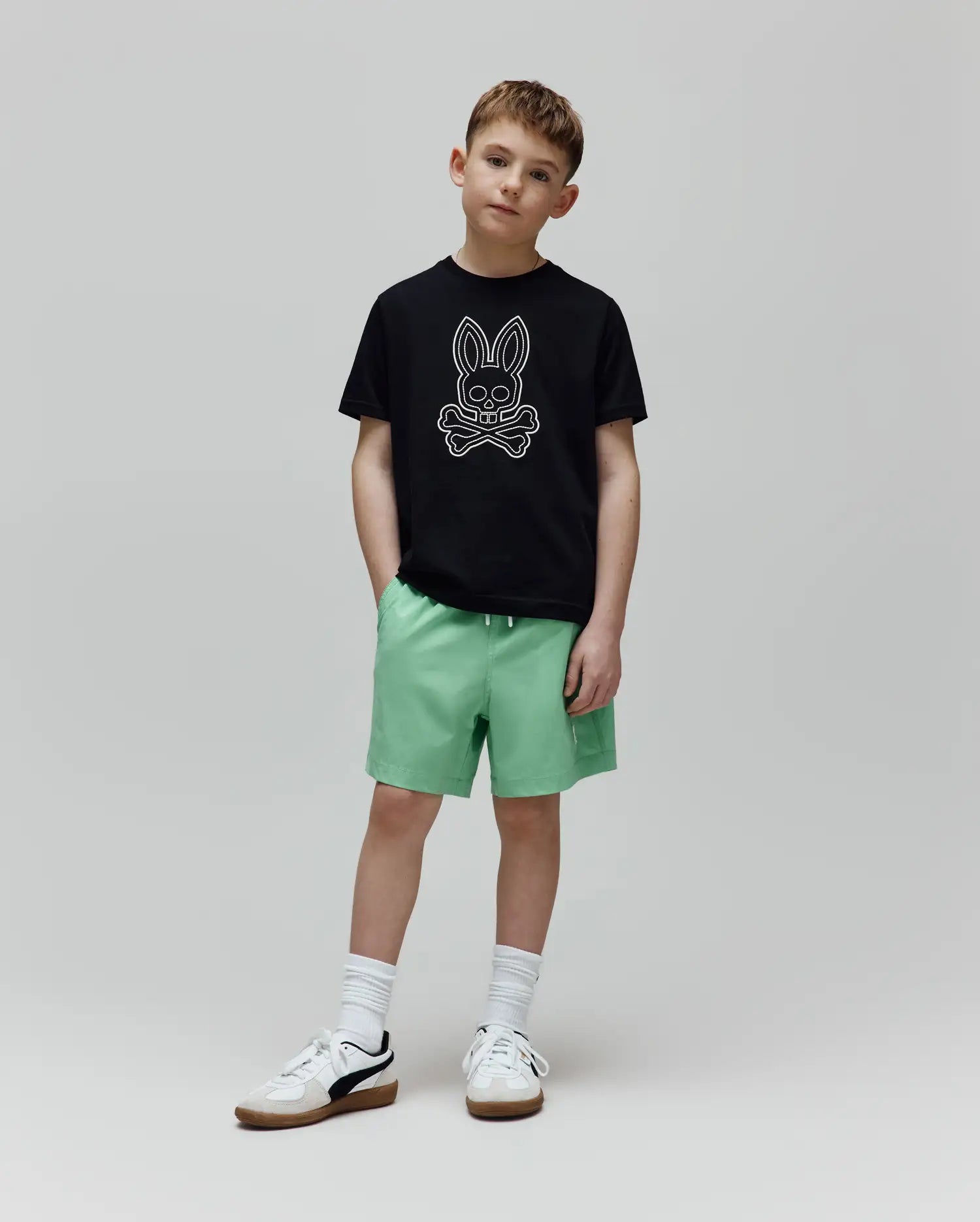 Black classic-fit jersey t-shirt with a large white outline Psycho Bunny skull logo on the chest, and light green Kids Samos Hydrochromic Printed Swim Trunk with a white drawstring.