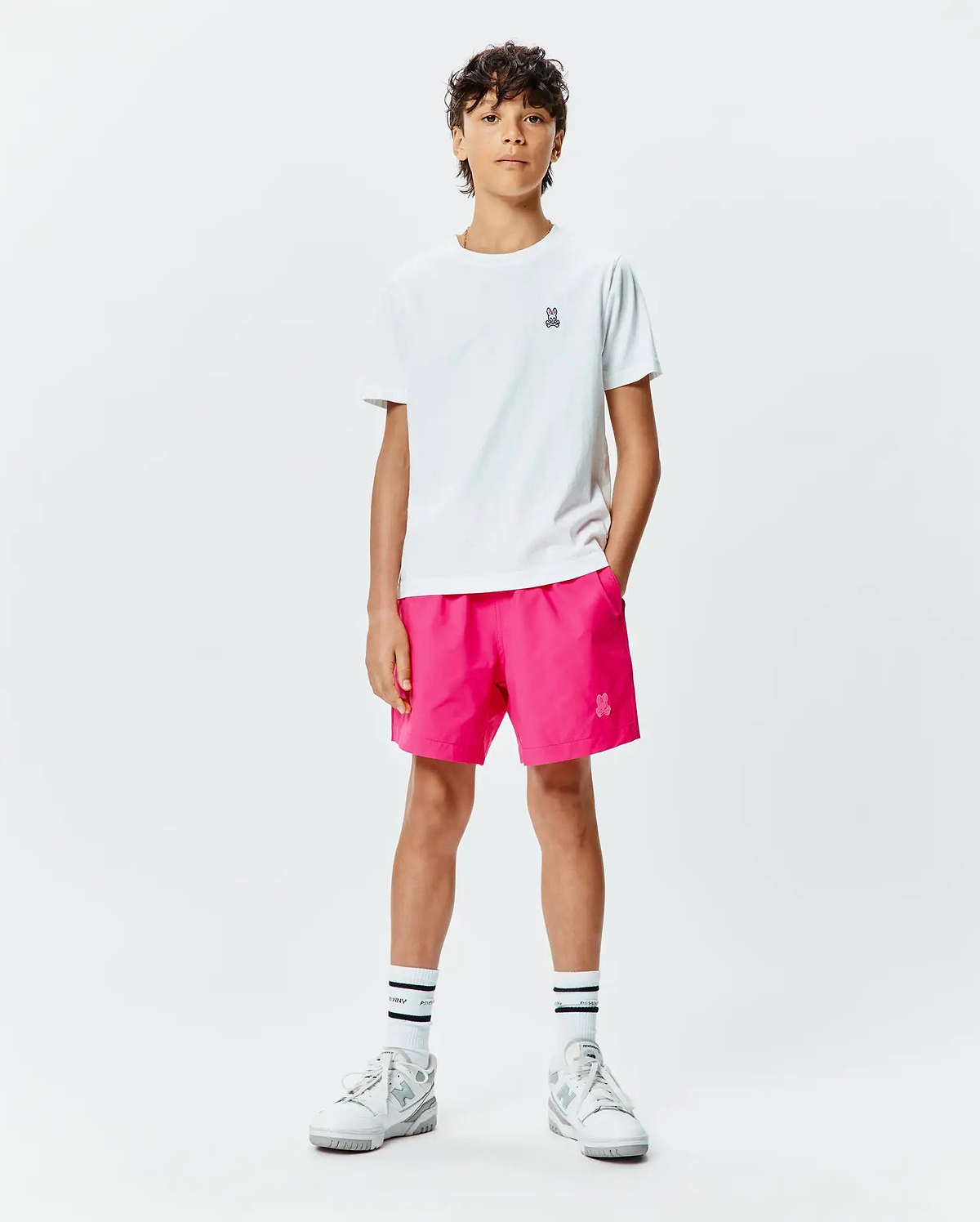 White classic fit jersey t-shirt with a small embroidered Psycho Bunny skull logo on the left chest, paired with hot pink KIDS SAMOS HYDROCHROMIC PRINTED SWIM TRUNK featuring a small embroidered bunny logo on the left thigh. (5)