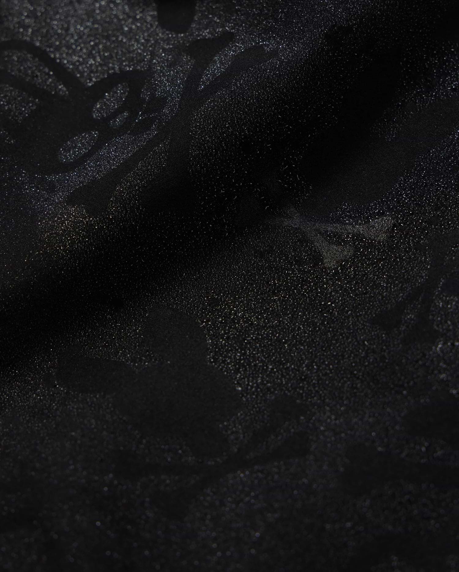 Close-up of Psycho Bunny's KIDS PARKER HYDROCHROMIC SWIM TRUNK (B0W646C200) featuring a subtle, intricate pattern of skulls, bones, and abstract shapes that creates a textured, shadowy effect on the black fabric. The quick-dry material boasts a glossy finish with a slightly shimmering surface.