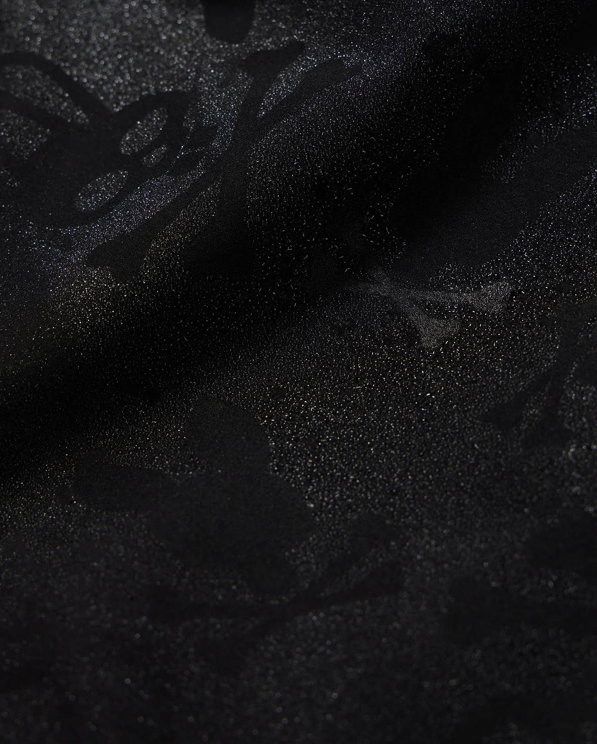 Close-up of Psycho Bunny's KIDS PARKER HYDROCHROMIC SWIM TRUNK (B0W646C200) featuring a subtle, intricate pattern of skulls, bones, and abstract shapes that creates a textured, shadowy effect on the black fabric. The quick-dry material boasts a glossy finish with a slightly shimmering surface.