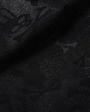 Close-up of Psycho Bunny's KIDS PARKER HYDROCHROMIC SWIM TRUNK (B0W646C200) featuring a subtle, intricate pattern of skulls, bones, and abstract shapes that creates a textured, shadowy effect on the black fabric. The quick-dry material boasts a glossy finish with a slightly shimmering surface.