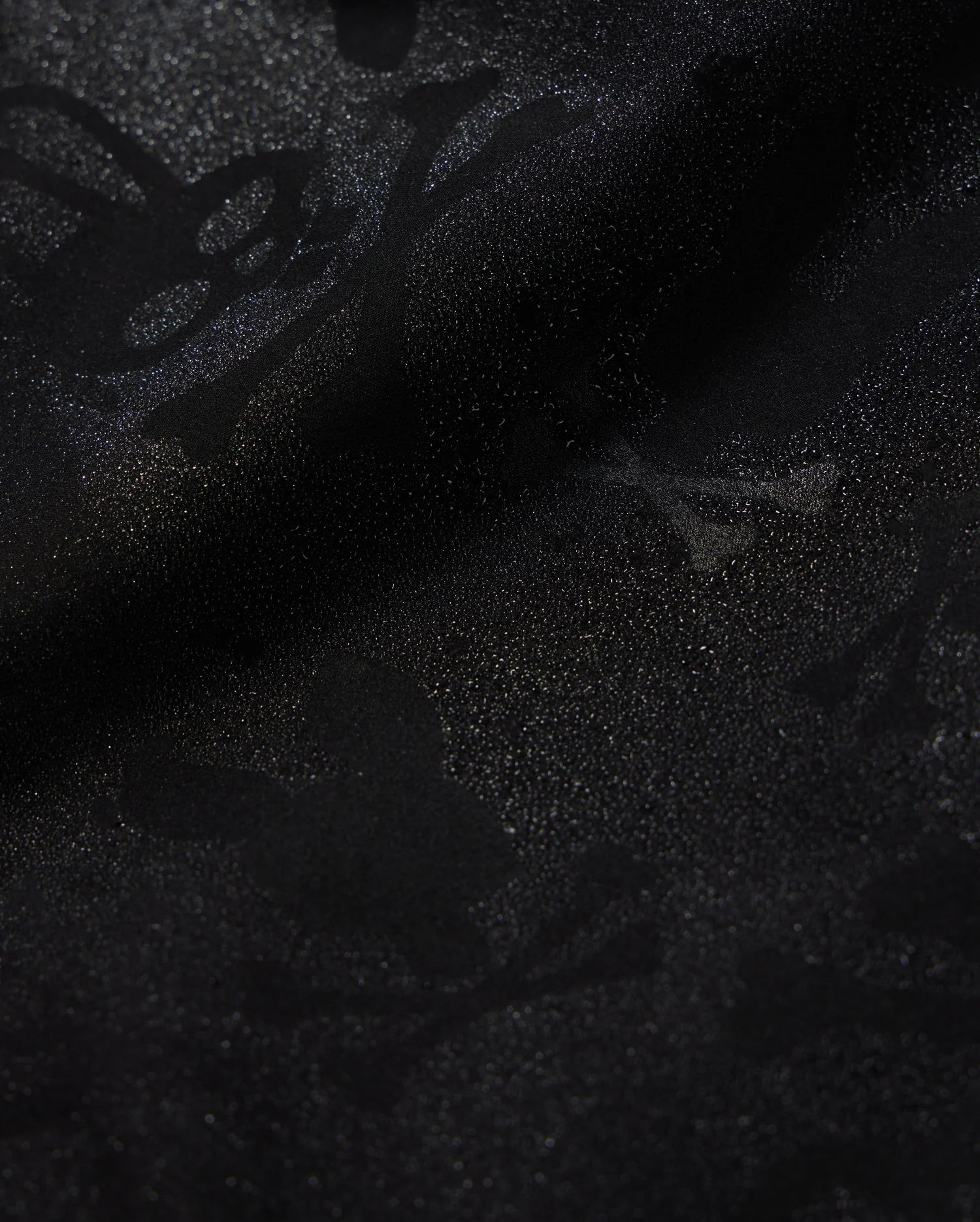 Close-up of Psycho Bunny's KIDS PARKER HYDROCHROMIC SWIM TRUNK (B0W646C200) featuring a subtle, intricate pattern of skulls, bones, and abstract shapes that creates a textured, shadowy effect on the black fabric. The quick-dry material boasts a glossy finish with a slightly shimmering surface.