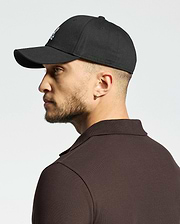 A man wears a Psycho Bunny MENS CLASSIC BASEBALL CAP (B6A816A1HT) in black, perfectly paired with a dark polo shirt. He sports a stud earring in his left ear, gazing sideways against a light background.