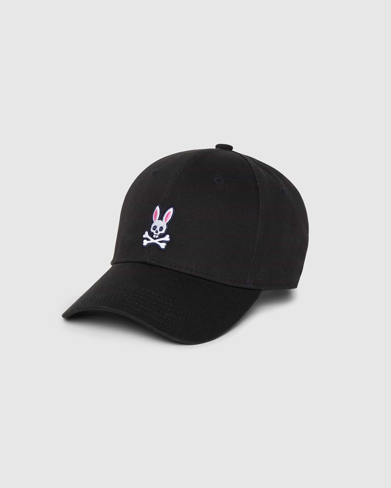 The Psycho Bunny Men's Classic Baseball Cap (B6A816A1HT) is crafted from 100% cotton in classic black, featuring their iconic pink and white embroidered Bunny logo with crossbones on the front. This cap includes a curved brim, six panels with eyelets for ventilation, all set against a simple white background.