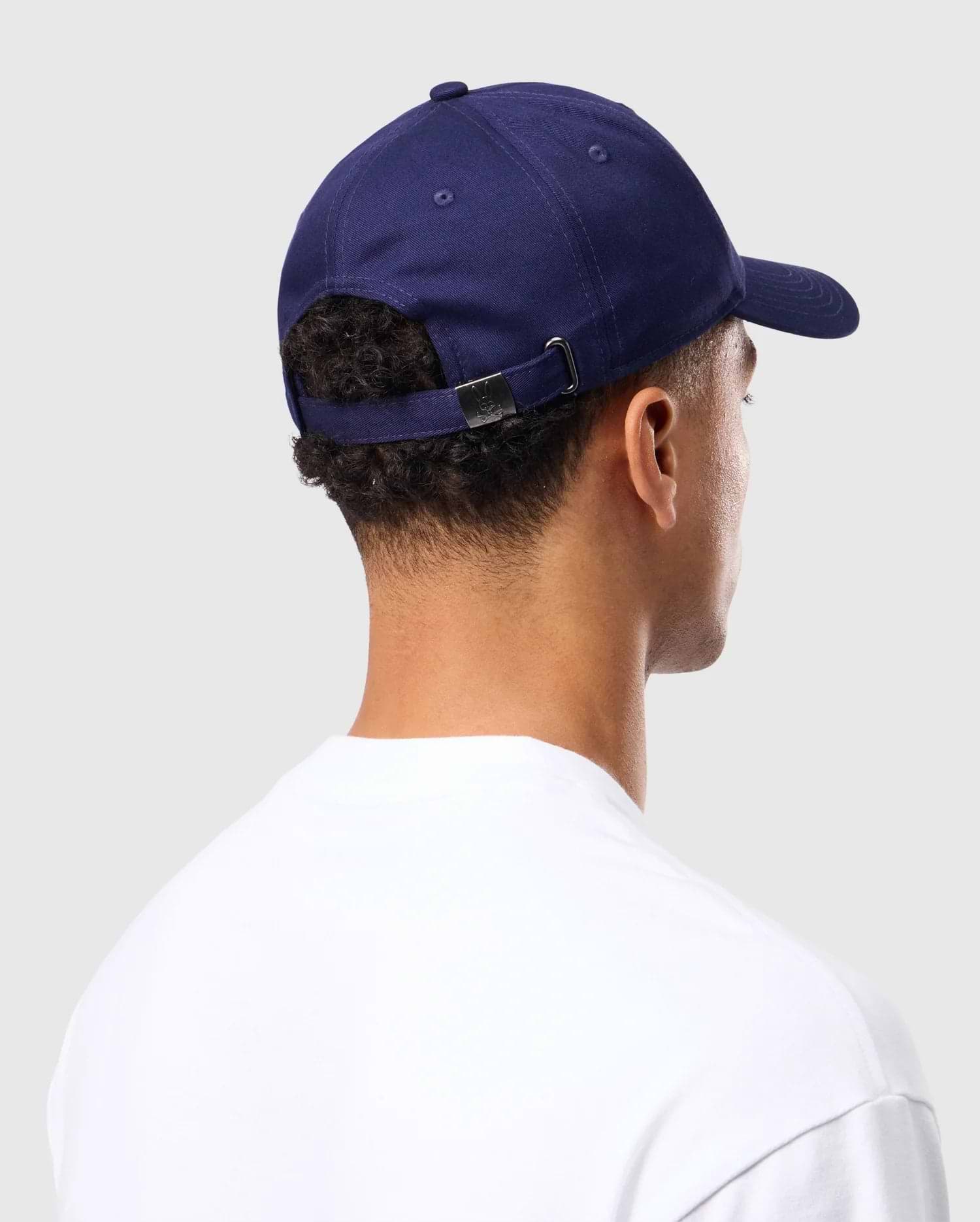 A person in a classic white shirt faces away, wearing the MENS CLASSIC BASEBALL CAP - B6A816A1HT by Psycho Bunny, which is a navy blue six-panel cotton twill cap with an adjustable strap. The background is plain light gray.
