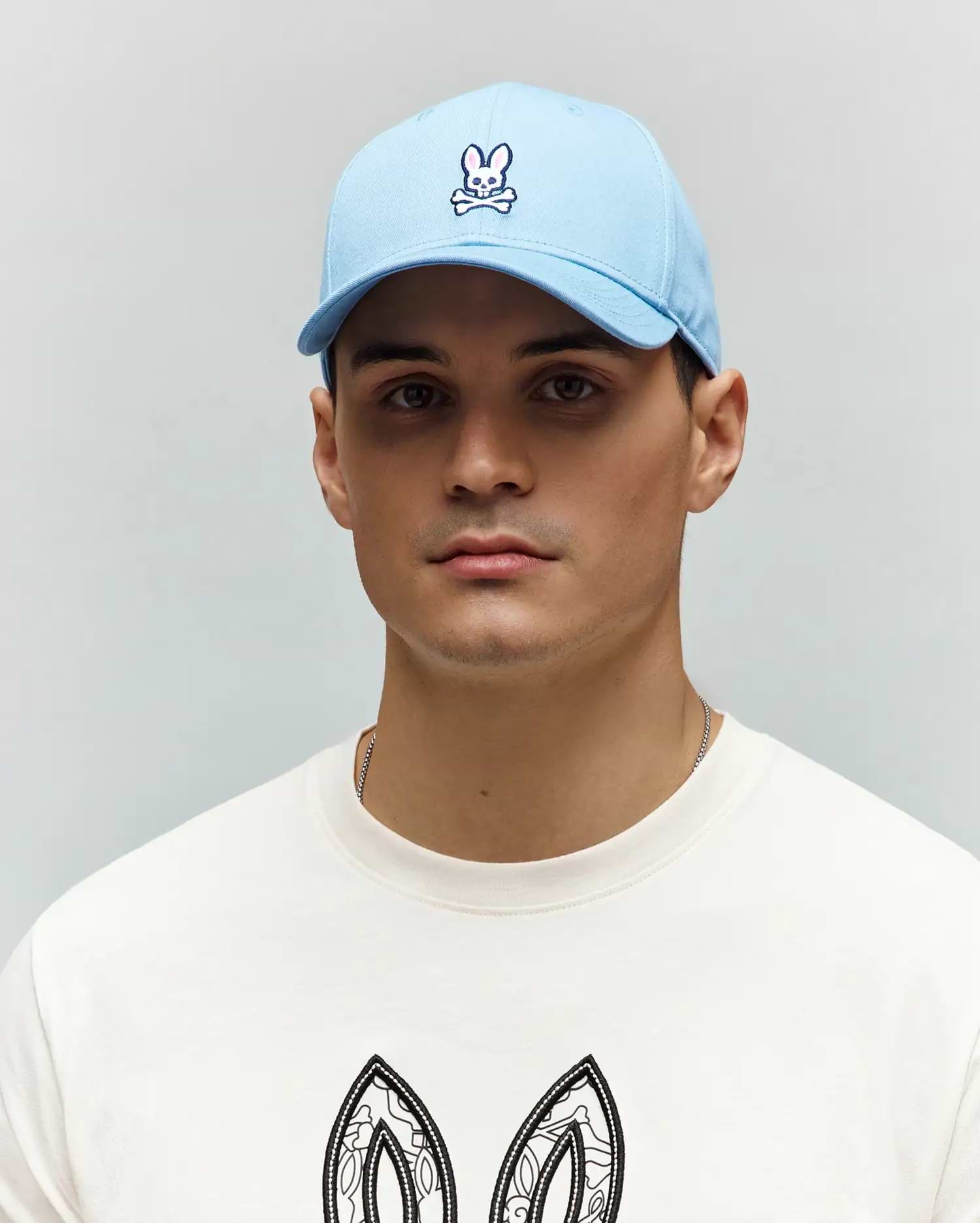 Mens Classic Baseball Cap in sky blue, featuring a prominent embroidered white and navy Psycho Bunny skull logo, alongside an off-white jersey t-shirt with a large, intricate black and white bunny ear graphic.