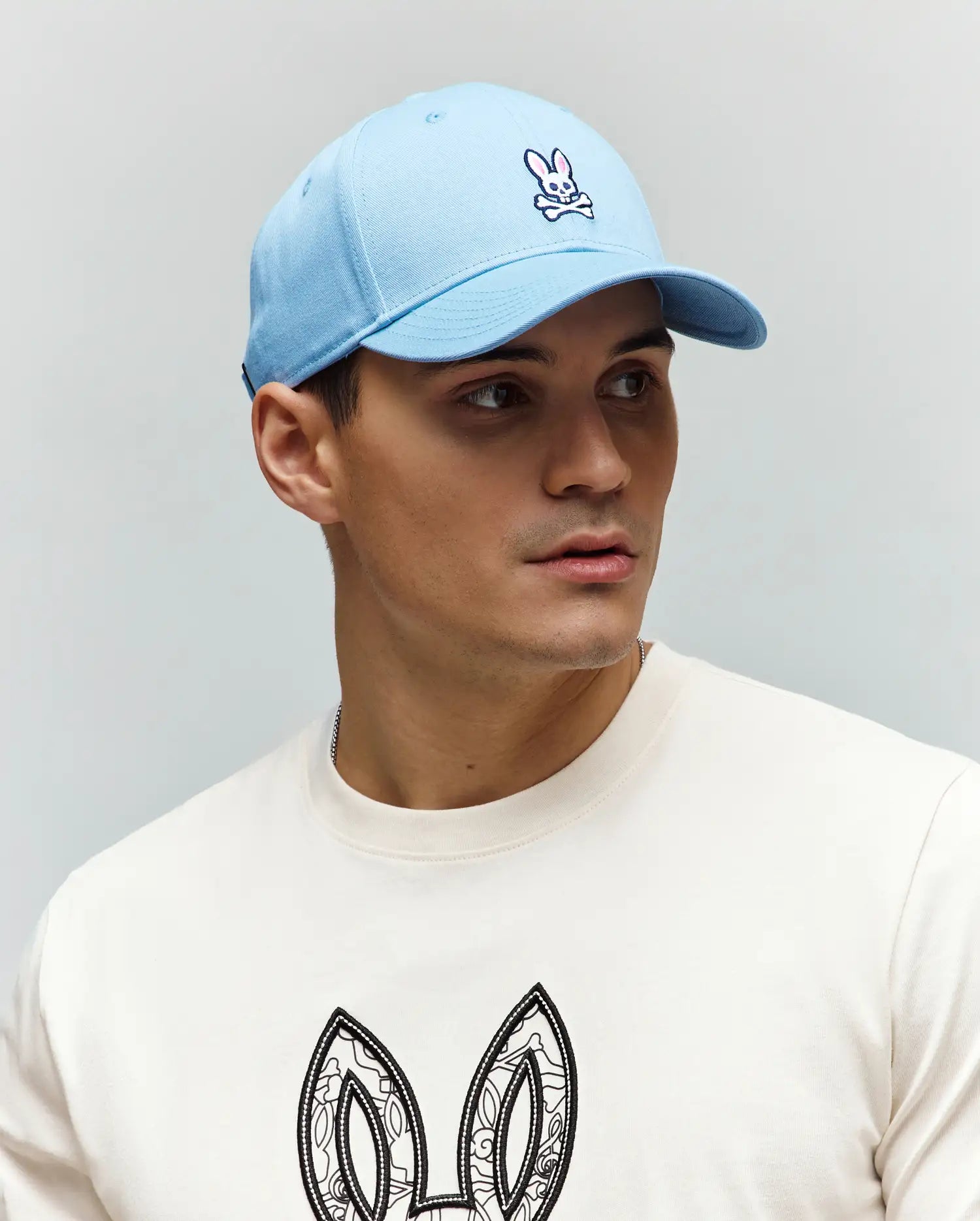 Light blue twill Mens Classic Baseball Cap with embroidered pink and white Psycho Bunny skull logo. Off-white jersey crewneck t-shirt features large, intricate black and white bunny ears graphic.