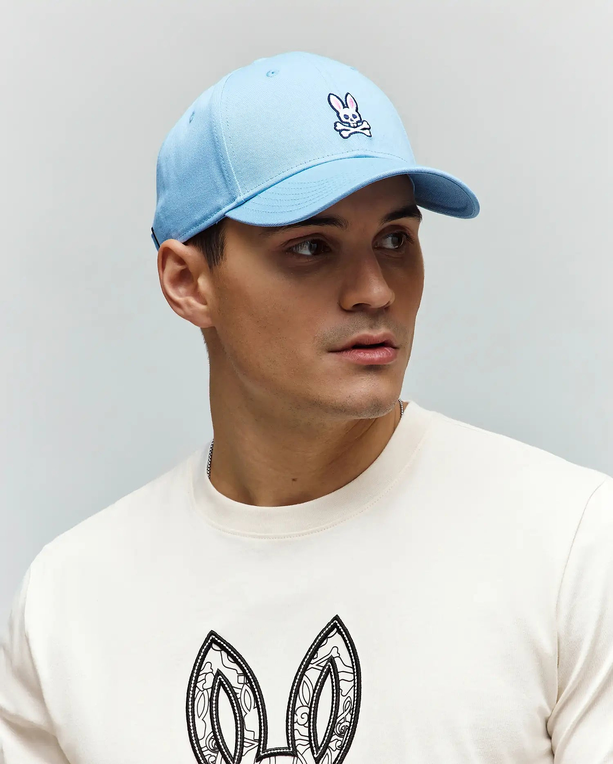Light blue twill Mens Classic Baseball Cap with embroidered pink and white Psycho Bunny skull logo. Off-white jersey crewneck t-shirt features large, intricate black and white bunny ears graphic.