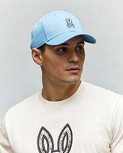 Light blue twill Mens Classic Baseball Cap with embroidered pink and white Psycho Bunny skull logo. Off-white jersey crewneck t-shirt features large, intricate black and white bunny ears graphic.