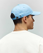 Mens Classic Baseball Cap in light blue cotton twill, featuring an adjustable strap with a small dark metal Psycho Bunny skull logo buckle, paired with a white jersey t-shirt.