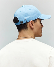 Mens Classic Baseball Cap in light blue cotton twill, featuring an adjustable strap with a small dark metal Psycho Bunny skull logo buckle, paired with a white jersey t-shirt.