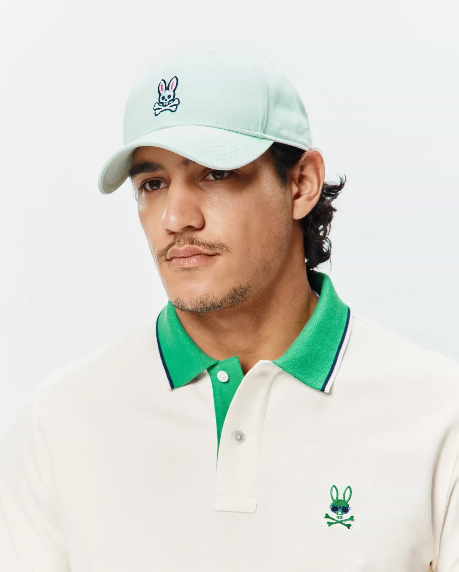 Cream pique polo with green collar, navy stripe, and embroidered Psycho Bunny logo with sunglasses, paired with a mint Mens Classic Baseball Cap featuring an embroidered bunny skull.
