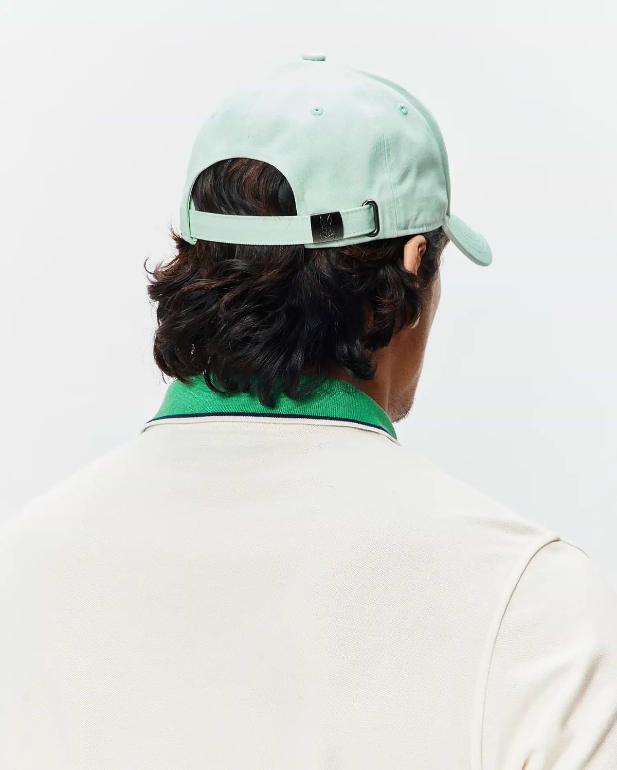 Mint Green Mens Classic Baseball Cap with a small Psycho Bunny skull logo on the metal buckle, paired with a cream pique polo featuring a green ribbed collar and thin navy stripe.