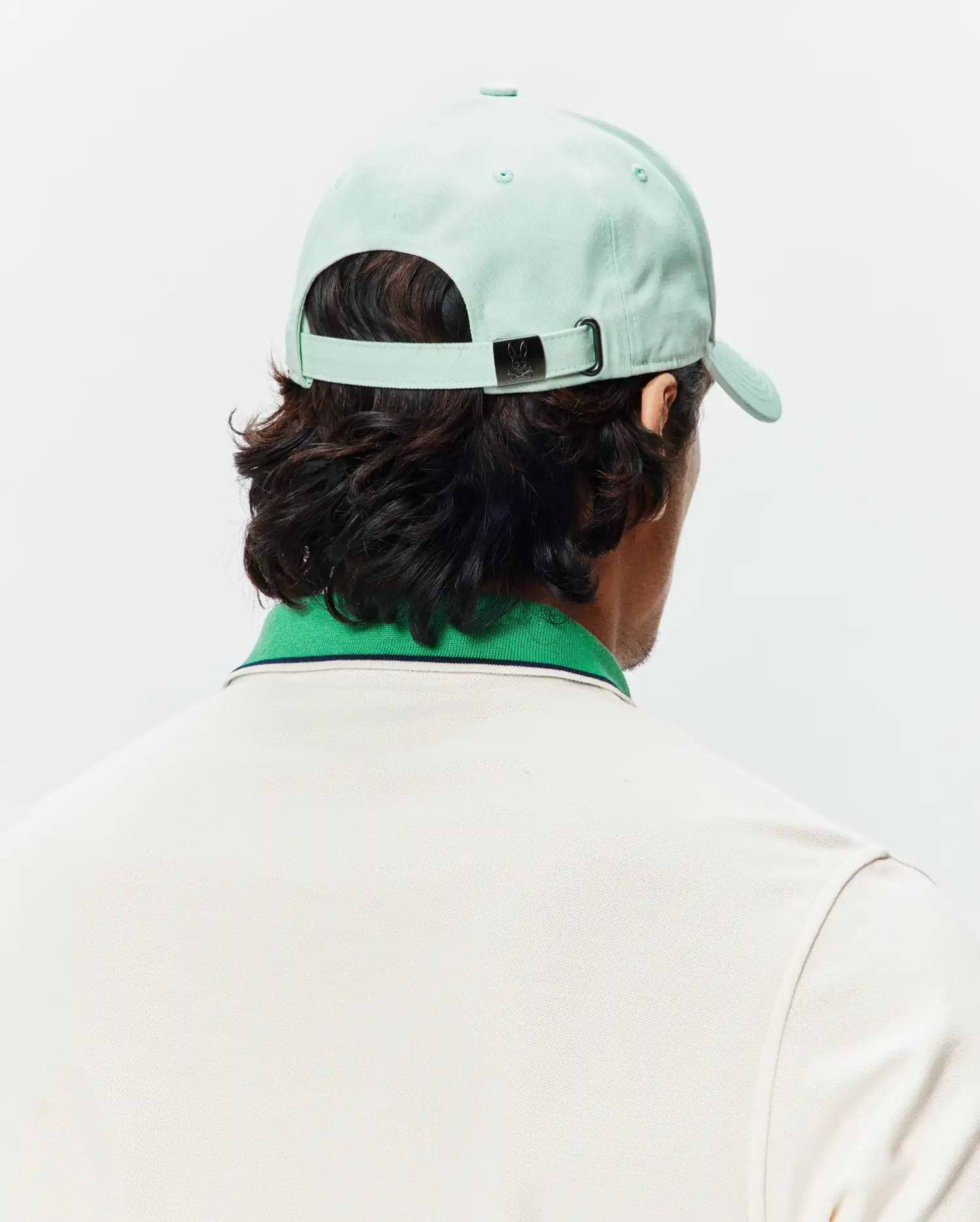 Mint Green Mens Classic Baseball Cap with a small Psycho Bunny skull logo on the metal buckle, paired with a cream pique polo featuring a green ribbed collar and thin navy stripe.
