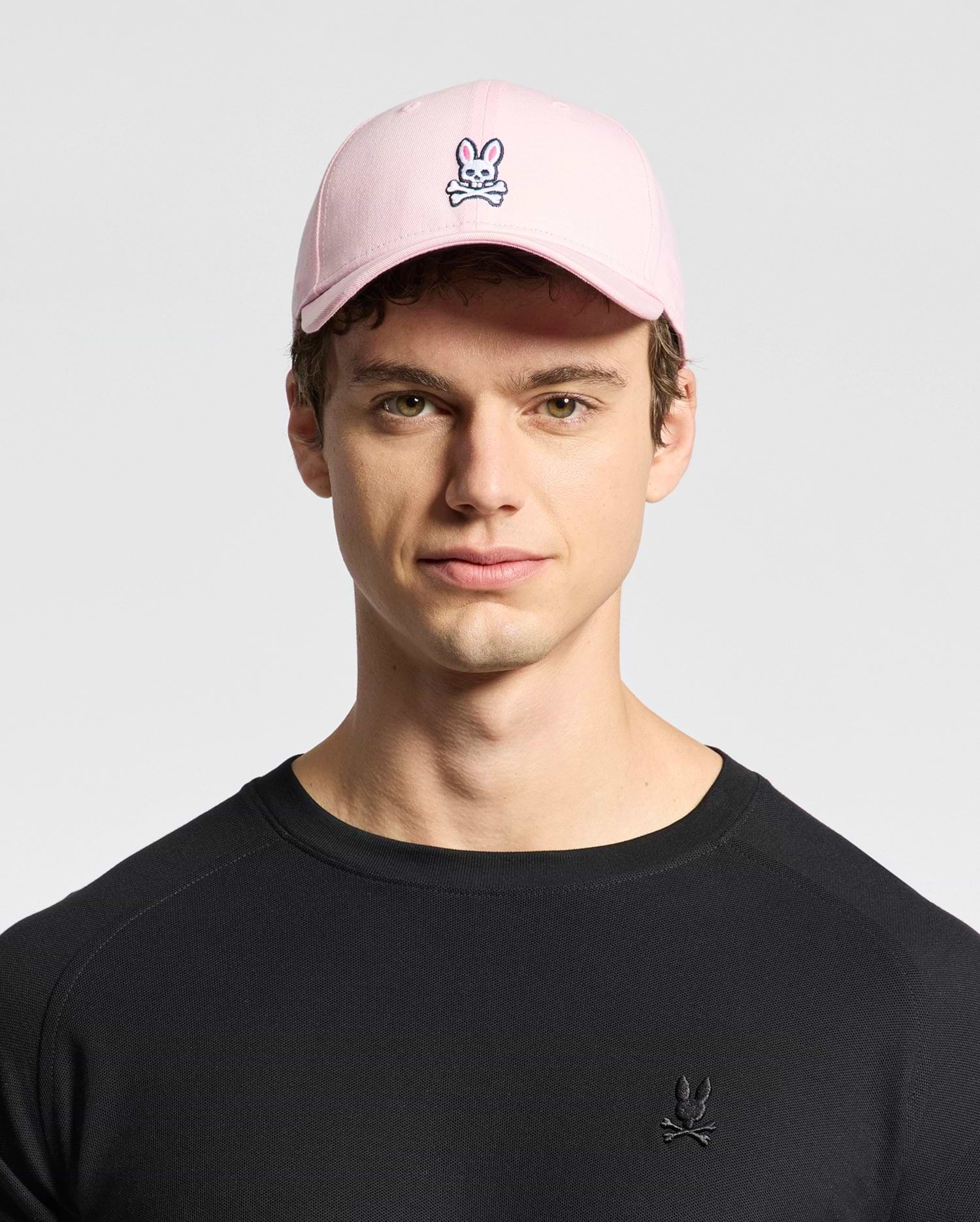 A young person sports Psycho Bunny's MENS CLASSIC BASEBALL CAP - B6A816A1HT made of cotton twill with an embroidered bunny and crossbones logo. Their black shirt features the same playful design, standing out against the plain background.