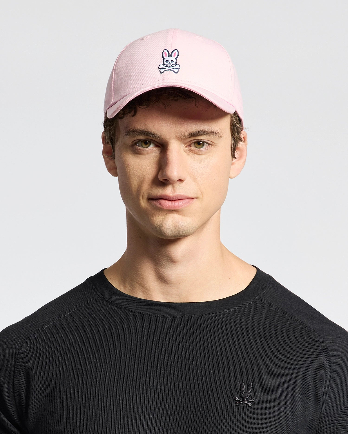 A young person sports Psycho Bunny's MENS CLASSIC BASEBALL CAP - B6A816A1HT made of cotton twill with an embroidered bunny and crossbones logo. Their black shirt features the same playful design, standing out against the plain background.