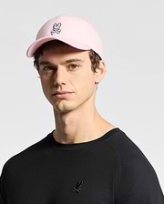 Wearing a pink cotton twill MENS CLASSIC BASEBALL CAP - B6A816A1HT by Psycho Bunny with an embroidered Bunny logo, and a black shirt with a small logo, the person maintains a neutral expression while facing the camera against a light background.
