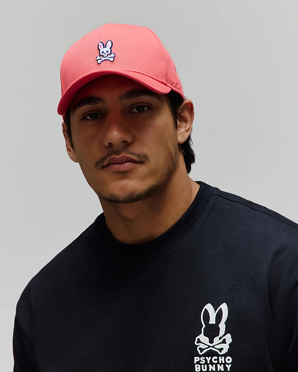 Mens Classic Baseball Cap in coral with an embroidered Psycho Bunny skull logo, and a black jersey crewneck sweatshirt featuring a large white embroidered Psycho Bunny logo and text.