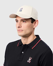 A person is stylishly dressed in a Psycho Bunny ensemble—a beige MENS CLASSIC BASEBALL CAP - B6A816B200, 100% cotton, and a black polo shirt. Both feature the rabbit graphic. The subtle gray background enhances the casually elegant look.