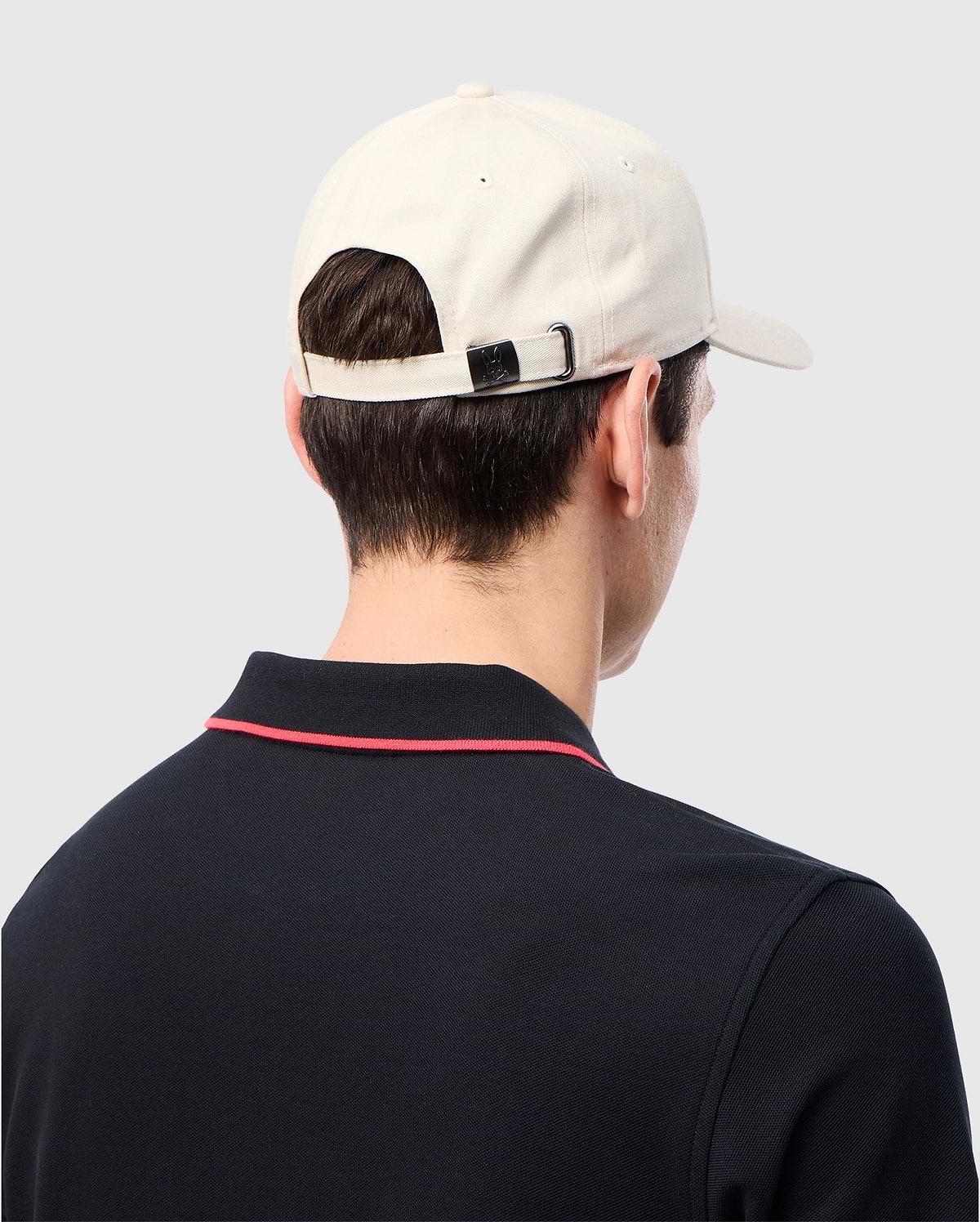 A man wearing a Psycho Bunny MENS CLASSIC BASEBALL CAP - B6A816B200 in beige and a black collared shirt with red trim stands backwards, highlighting the cap and shirt's design details.