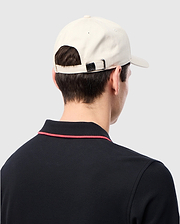 A man wearing a Psycho Bunny MENS CLASSIC BASEBALL CAP - B6A816B200 in beige and a black collared shirt with red trim stands backwards, highlighting the cap and shirt's design details.