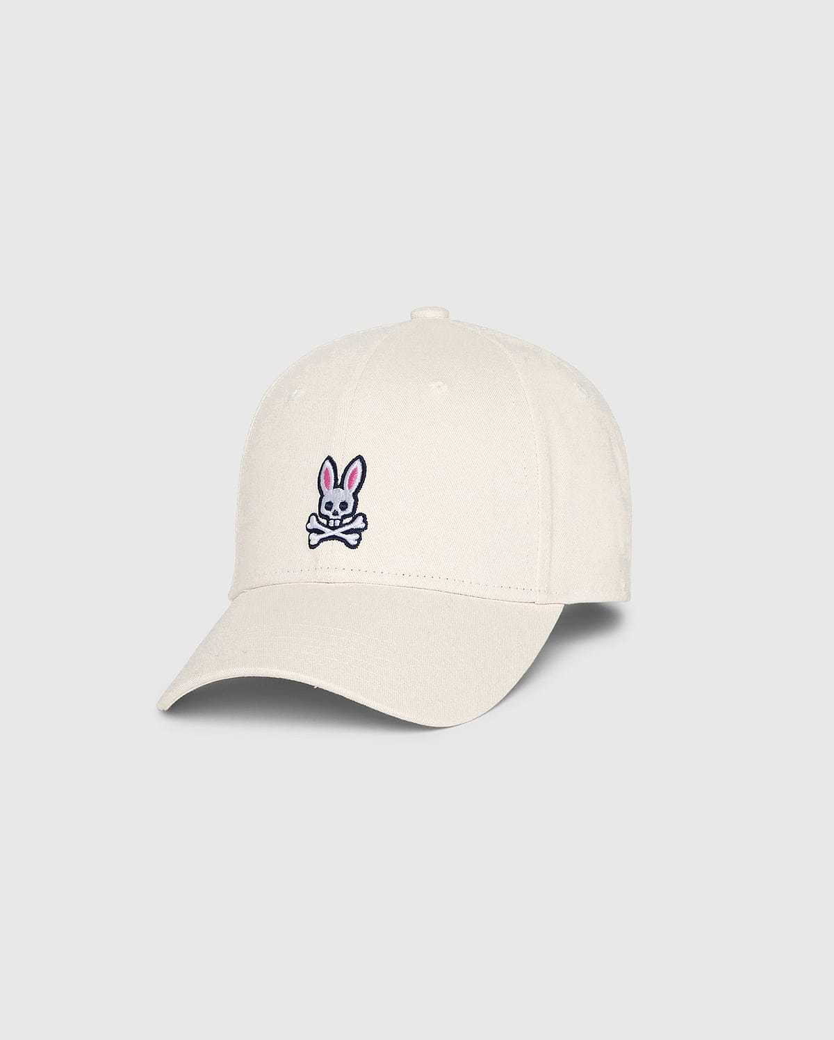 The Psycho Bunny MENS CLASSIC BASEBALL CAP (B6A816B200), made from 100% cotton in beige, showcases a small pink bunny with crossed bones on the front. Angled to highlight both front and side, this standout accessory adds character to any collection.
