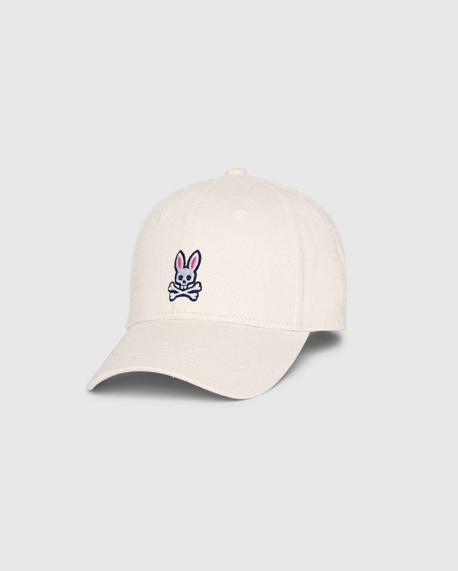 The Psycho Bunny MENS CLASSIC BASEBALL CAP (B6A816B200), made from 100% cotton in beige, showcases a small pink bunny with crossed bones on the front. Angled to highlight both front and side, this standout accessory adds character to any collection.